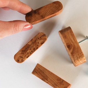 Könnte beinhalten: Vier Holzknaufgriffe mit natürlicher Holzoptik. Die Griffe sind rechteckig und haben eine leicht abgerundete Kante. Zwei der Griffe haben ein Schraubenloch in der Mitte. Die anderen beiden Griffe sind schlicht.