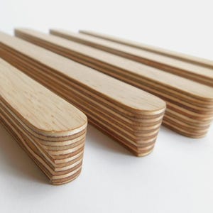 Peut inclure: Cinq patères murales en bois, en contreplaqué, avec une finition bois naturel. Les patères sont disposées en rangée, la plus proche étant en focus. Chaque patère a un bord arrondi et est composée de plusieurs couches de bois.