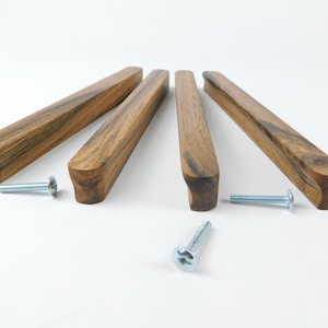 Könnte beinhalten: Vier Holzschrankgriffe mit natürlicher Holzmaserung. Jeder Griff ist rechteckig mit abgerundeten Kanten und einem gebogenen Ende. Silberschrauben sind für die Installation enthalten. Die Griffe sind auf einer weißen Oberfläche angeordnet.