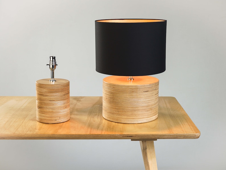 Puede incluir: Dos l&aacute;mparas de mesa de madera con pantallas negras. La l&aacute;mpara m&aacute;s grande tiene una base cil&iacute;ndrica hecha de chapa de madera apilada. La l&aacute;mpara m&aacute;s peque&ntilde;a tiene una base similar pero es m&aacute;s corta. Ambas l&aacute;mparas est&aacute;n sobre una mesa de madera.