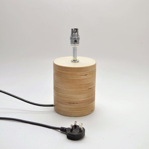 Puede incluir: Una l&aacute;mpara de mesa de madera con un cable negro y un enchufe brit&aacute;nico. La l&aacute;mpara tiene una base cil&iacute;ndrica de madera contrachapada y un portal&aacute;mparas cromado.