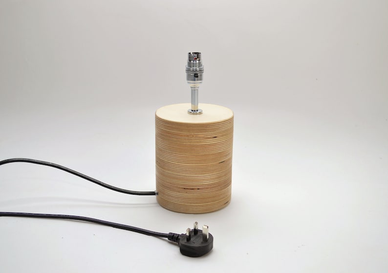 Puede incluir: Una l&aacute;mpara de mesa de madera con un cable negro y un enchufe brit&aacute;nico. La l&aacute;mpara tiene una base cil&iacute;ndrica de madera contrachapada y un portal&aacute;mparas cromado.