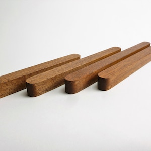 Könnte beinhalten: Vier rechteckige Holzstücke mit abgerundeten Enden sind auf einer weißen Oberfläche angeordnet. Das Holz hat einen warmen, rötlich-braunen Ton, und die Stücke sind von unterschiedlicher Länge. Die Objekte scheinen handgefertigt zu sein.