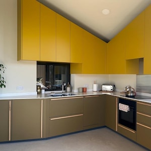 Könnte beinhalten: Eine moderne Küche mit gelben Oberschränken und braunen Unterschränken. Die Küche hat eine weiße Arbeitsplatte und ein Edelstahlspülbecken. Es gibt einen schwarzen Backofen und eine weiße Arbeitsplatte.