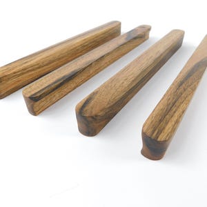 Könnte beinhalten: Vier Holz-Schubladengriffe mit einem natürlichen Holzmaserungsmuster. Die Griffe sind rechteckig mit abgerundeten Enden und diagonal auf weißem Hintergrund angeordnet. Das Holz hat eine Mischung aus hellen und dunklen Brauntönen.