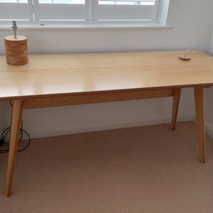 May include: A light wood desk with a simple design. The desk has a rectangular top and four legs. The desk is made of light wood and has a natural finish.