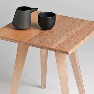 May include: A small wooden table with four legs and a top made of two pieces of plywood. The top is a light brown colour and has a natural wood grain. The table is decorated with a black ceramic jug and a black ceramic cup.