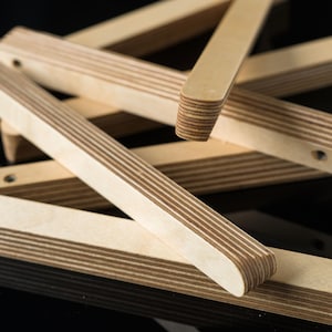 Könnte beinhalten: Ein Satz Holzlatten, jede mit einer mehrschichtigen Sperrholzkonstruktion, sind übereinander gestapelt und in einem Kreuzmuster angeordnet. Die Latten sind hellbraun und haben eine glatte, polierte Oberfläche.