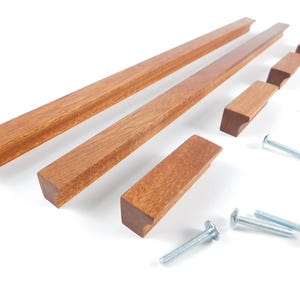 Könnte beinhalten: Eine Auswahl an Holzschrankgriffen und -griffen in verschiedenen Größen mit warmbraunem Finish. Die Hardware umfasst rechteckige Griffe und kleinere, quadratisch endende Griffe sowie silberne Schrauben zur Montage. Die Artikel sind auf einer weißen Oberfläche angeordnet.