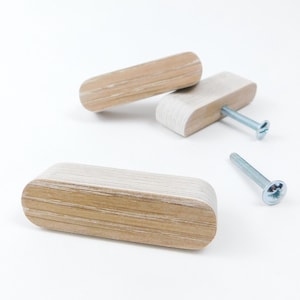 May include: Light wood cabinet pulls with a rounded rectangular design. Two pulls have screws inserted, and one screw is separate. The pulls are made of layered wood and have a natural finish. The cabinet pulls are made of layered wood.