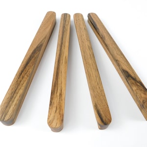 Puede incluir: Cuatro tiradores de cajón de madera con un patrón de vetas naturales. Los tiradores son rectangulares con extremos redondeados y están dispuestos sobre un fondo blanco. La madera tiene un color marrón cálido con vetas más oscuras.