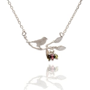 May include: A silver necklace with a delicate bird perched on a branch with leaves. The branch has a cluster of pink and green gemstones hanging from it.