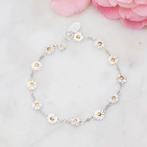 Könnte beinhalten: Ein silbernes Armband mit einer zarten Kette und kleinen Gänseblümchen-Anhängern. Die Gänseblümchen-Mittelpunkte sind golden.