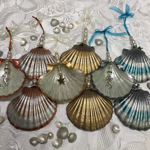 May include: Seven seashell ornaments painted in silver, gold, and teal. Each seashell has a pearl and charm hanging from it. The charms include a starfish, a seahorse, and a swirl.