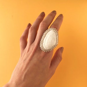 Puede incluir: Un anillo de concha blanca con un borde de cuentas. El anillo está en un dedo y el fondo es amarillo.