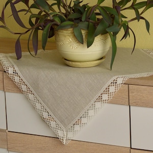 Può includere: Una pianta da appartamento in un vaso color crema con un piattino poggia su una tovaglietta beige con bordi in pizzo. La pianta ha foglie verdi e viola. La tovaglietta poggia su un mobile chiaro in legno e bianco con maniglie argentate.