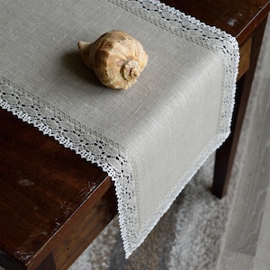 Puede incluir: Un camino de mesa de lino beige con ribete de encaje blanco, exhibido sobre una mesa de madera oscura. Una concha marina descansa sobre el camino de mesa. El camino de mesa añade elegancia.