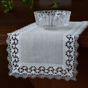 Puede incluir: Un camino de mesa de lino blanco con un delicado borde de encaje. El camino de mesa está sobre una mesa de madera oscura con un cuenco de vidrio transparente en el centro.