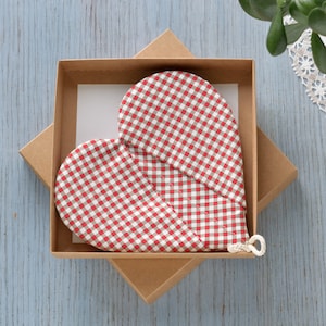 Puede incluir: Un guante de cocina en forma de corazón con un patrón de cuadros rojos y blancos, presentado en una caja de regalo de cartón marrón. El guante tiene un lazo para colgar. La caja está abierta, revelando el guante y una tarjeta blanca.