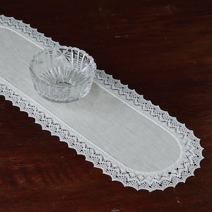 Op de afbeelding: Een witte ovale tafelloper met een delicate kanten rand en een heldere, geslepen glazen kom. De loper ligt op een donker houten oppervlak, wat een contrast creëert.