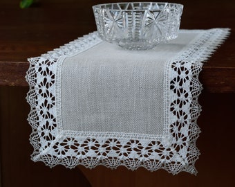 Puede incluir: Un camino de mesa de lino blanco con un delicado borde de encaje. El camino de mesa está sobre una mesa de madera oscura con un cuenco de vidrio transparente en el centro.