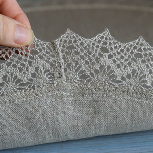 May include: A round, beige linen tablecloth with a delicate crocheted lace trim. The lace is a light beige color and has a floral pattern.