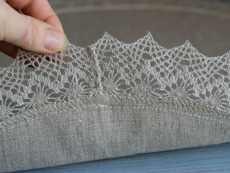 May include: A round, beige linen tablecloth with a delicate crocheted lace trim. The lace is a light beige color and has a floral pattern.