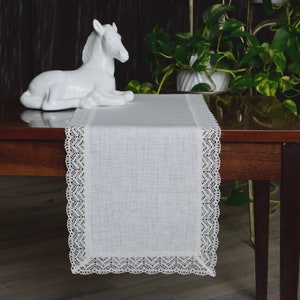 Può includere: Runner da tavolo in lino bianco con bordatura in pizzo color crema. Il runner rettangolare è appoggiato su un tavolo in legno scuro. Una statuina di cavallo in ceramica bianca e piante in vaso sono sullo sfondo.