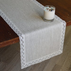 Op de afbeelding: Een witte linnen tafelrunner met een witte kantversiering. De runner ligt op een houten tafel met een kaars in een glazen pot.