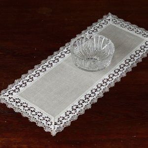 Puede incluir: Un camino de mesa de encaje blanco con un cuenco de vidrio transparente en el centro. El camino de mesa está sobre una superficie de madera marrón oscura.