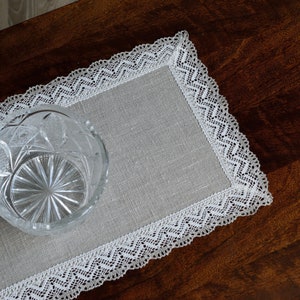Pode incluir: Um  placemat de renda branca com um centro de linho cinza. O placemat está sobre uma mesa de madeira.