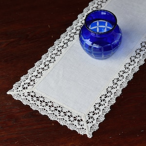 May include: A white linen table runner with delicate lace trim, featuring a cobalt blue glass vase. The runner is rectangular and rests on a dark wooden surface, creating a contrast of textures and colors. The vase has a geometric pattern.