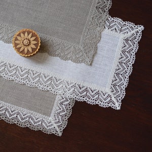 Può includere: Due tovagliette quadrate in lino con un delicato bordo in pizzo bianco. La tovaglietta superiore è di colore grigio chiaro e quella inferiore è bianca. Una piccola scatola decorativa in legno con un motivo floreale si trova sopra le tovagliette.