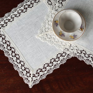 Könnte beinhalten: Zwei weiße Tischsets aus Leinen mit zarter Spitzenborte. Eine weiße Teetasse und ein Unterteller mit Blumenmuster liegen auf den Tischsets.