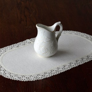 May include: White oval table runner with a delicate lace border. A white ceramic jug with a floral design sits on the runner.
