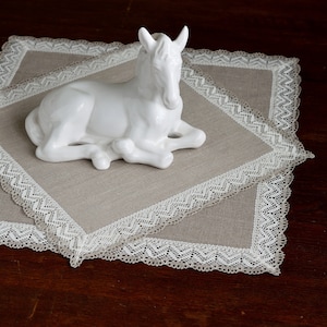 Könnte beinhalten: Eine weiße Porzellan-Pferdefigur, die auf einem beigefarbenen quadratischen Platzdeckchen aus Leinen mit weißer Spitzenborte liegt.