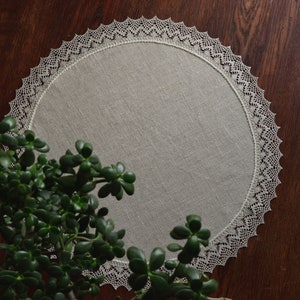 May include: A round, white linen table runner with a delicate lace border. The runner is on a dark wood surface with a green plant in the foreground.