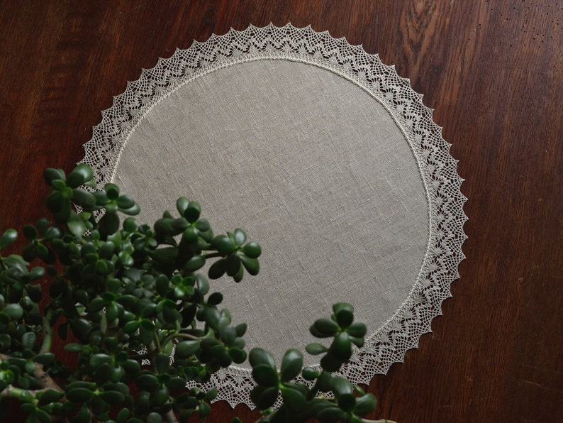 May include: A round, white linen table runner with a delicate lace border. The runner is on a dark wood surface with a green plant in the foreground.