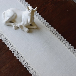 Könnte beinhalten: Ein weißer Tischläufer aus Leinen mit einer zarten Spitzenborte. Eine weiße Keramikfigur eines Pferdes liegt auf dem Läufer.