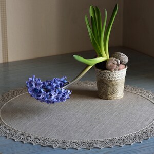 May include: A blue hyacinth flower in bloom, with green leaves, is displayed in a small, decorative pot with a lace trim. The pot is sitting on a round, gray linen placemat with a lace border.