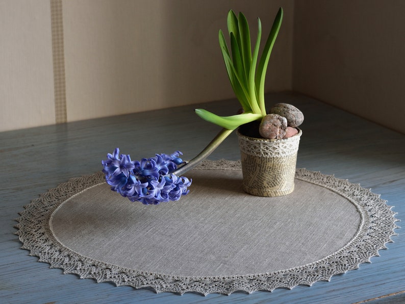 May include: A blue hyacinth flower in bloom, with green leaves, is displayed in a small, decorative pot with a lace trim. The pot is sitting on a round, gray linen placemat with a lace border.