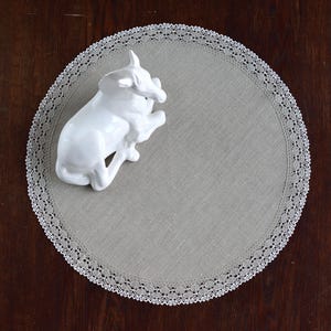 Op de afbeelding: Een witte keramische stierfiguur ligt op een ronde, lichtgrijze linnen placemat met een delicate witte kanten rand. De placemat staat op een donkerbruin houten oppervlak, wat een contrast in texturen en kleuren creëert.