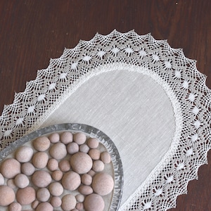 Puede incluir: Un camino de mesa ovalado de lino blanco con un delicado borde de encaje de ganchillo. El camino de mesa está sobre una superficie de madera oscura y tiene un cuenco decorativo de pequeñas piedras lisas marrones en el centro.