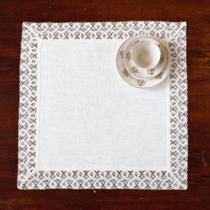 May include: White linen placemat with a white lace border. A small white teacup sits inside a floral patterned saucer.