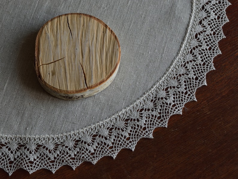 May include: A round wooden coaster with a natural wood grain finish and a light gray linen tablecloth with a delicate lace trim.
