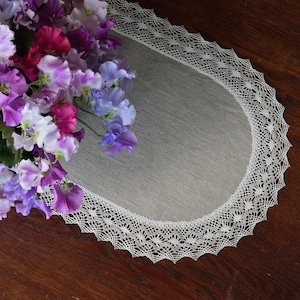 Può includere: Un runner da tavolo ovale bianco con un delicato bordo in pizzo. Il runner è in lino e ha una texture sottile. Un bouquet di piselli odorati viola, rosa e bianchi è posto sopra il runner.