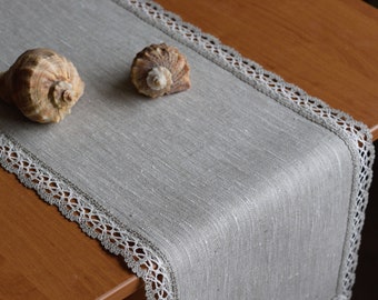 Puede incluir: Camino de mesa de lino gris con ribete de encaje blanco. Dos conchas marinas descansan sobre el camino de mesa. El camino de mesa está sobre una mesa de madera. Para la decoración del hogar.