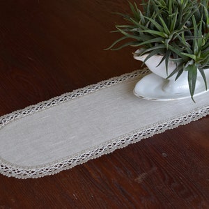 以下が含まれることがあります： 白のクロシェレースの縁取りが施されたベージュのリネン製テーブルランナー。ランナーは濃い木製のテーブルの上にあり、小さな白い鉢に緑の多肉植物が入っています。