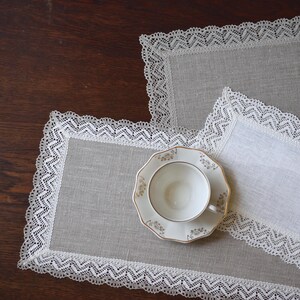 Könnte beinhalten: Nahaufnahme eines Teeservices auf einem Holztisch. Das Set umfasst eine weiße Teetasse und Untertasse mit goldenen Verzierungen, platziert auf einer Leinen-Tischdecke mit weißer Spitzenborte. Zwei weitere Tischsets mit Spitzenbesatz sind ebenfalls sichtbar.
