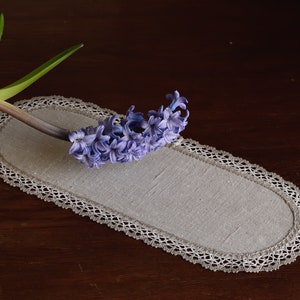 Puede incluir: Una flor de jacinto de color púrpura claro sobre un camino de mesa ovalado de lino blanco con borde de encaje. El camino de mesa está sobre una superficie de madera marrón oscura.